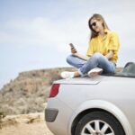 woman in yellow blouse and blue jeans taking selfie while sitting on car